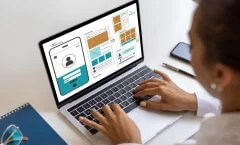 Uma pessoa trabalha em um notebook sobre uma mesa clara, desenvolvendo wireframes e protótipos de interface digital. Na tela, aparecem layouts de site e aplicativo mobile, com blocos que representam áreas de login, feed de conteúdo, galerias e seções de perfil de usuário.