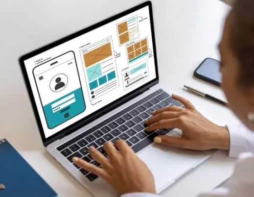 Uma pessoa trabalha em um notebook sobre uma mesa clara, desenvolvendo wireframes e protótipos de interface digital. Na tela, aparecem layouts de site e aplicativo mobile, com blocos que representam áreas de login, feed de conteúdo, galerias e seções de perfil de usuário.