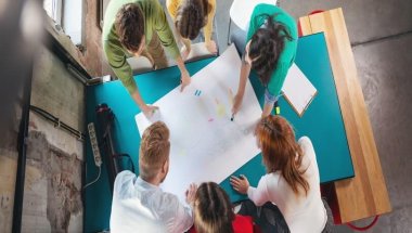 Um grupo multiétnico de pessoas está sentado ao redor de uma mesa de trabalho. No centro, está posicionada uma cartolina branca. Várias pessoas escrevem sobre ela, usando como facilitador a Metodologia Ágil.
