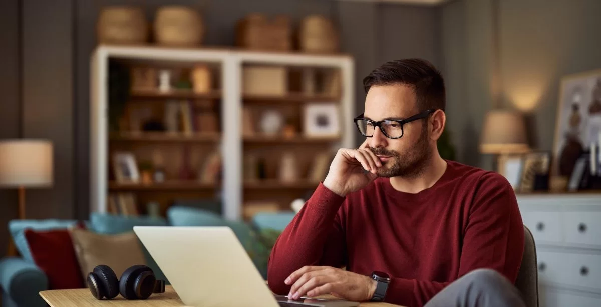 A imagem mostra um homem trabalhando no que parece ser a sala de estar de uma casa. Ele está olhando para o notebook com uma expressão pensativa, analisando os principais pontos entre GraphQL vs REST.
