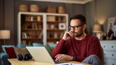 A imagem mostra um homem trabalhando no que parece ser a sala de estar de uma casa. Ele está olhando para o notebook com uma expressão pensativa, analisando os principais pontos entre GraphQL vs REST.