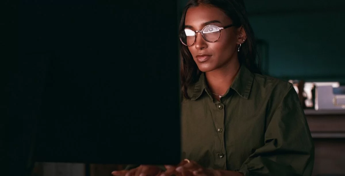 A imagem mostra uma desenvolvedora negra trabalhando na frente de um computador. Como o ambiente está escuro, pressupõe-se que ela esteja trabalhando no período noturno, possivelmente checando alguma API para desenvolvedores.
