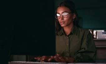 A imagem mostra uma desenvolvedora negra trabalhando na frente de um computador. Como o ambiente está escuro, pressupõe-se que ela esteja trabalhando no período noturno, possivelmente checando alguma API para desenvolvedores.