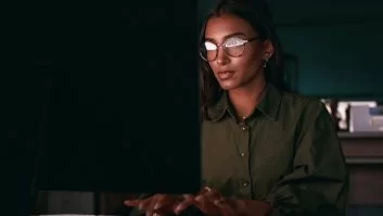 A imagem mostra uma desenvolvedora negra trabalhando na frente de um computador. Como o ambiente está escuro, pressupõe-se que ela esteja trabalhando no período noturno, possivelmente checando alguma API para desenvolvedores.