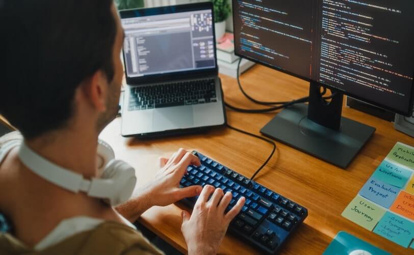 A imagem mostra um programador digitando em um teclado mecânico enquanto trabalha em um ambiente de desenvolvimento com dois monitores e um notebook exibindo linhas de código e interfaces de software. Sobre a mesa, há notas adesivas com termos como “UX Design”, “Wireframe” e “User Journey”, sugerindo o foco em experiência do usuário e design de interfaces. O cenário reflete a rotina moderna de um profissional de tecnologia, unindo programação, prototipagem e análise de sistemas. A iluminação quente e o ambiente organizado transmitem concentração, criatividade e produtividade, características típicas do universo tech e de desenvolvimento de aplicativos.