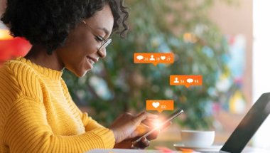 Mulher administrando as redes sociais on-line, sentada em um café dentro de casa. Na foto há colagem com ícones representando número de seguidores, curtidas e comentários em redes sociais.