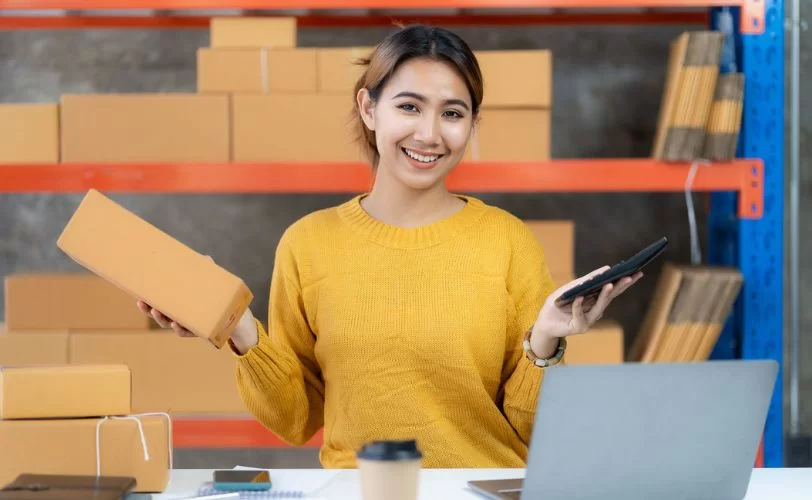 mulher com pacote e calculadora em mãos em frente ao notebook