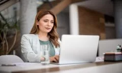 A imagem mostra uma mulher com roupas sociais usando um notebook no que parece ser um escritório. Ela aparenta ter um olhar confiante e seguro, pois está acessando um site que utiliza Wildcard SSL para aumentar a segurança dos dados.