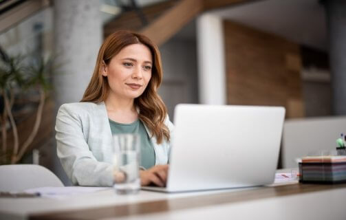 A imagem mostra uma mulher com roupas sociais usando um notebook no que parece ser um escritório. Ela aparenta ter um olhar confiante e seguro, pois está acessando um site que utiliza Wildcard SSL para aumentar a segurança dos dados.