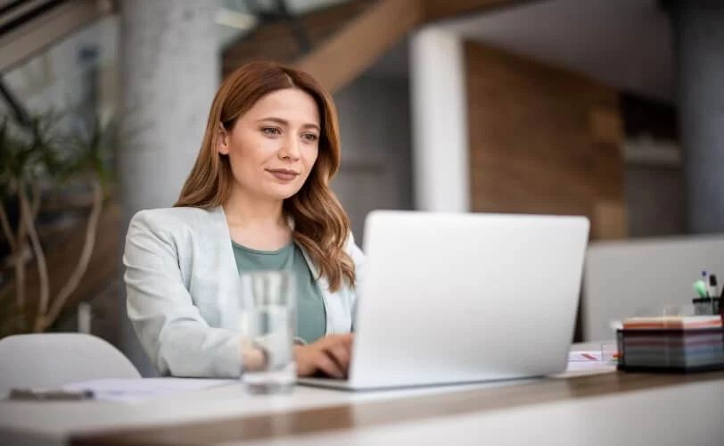 A imagem mostra uma mulher com roupas sociais usando um notebook no que parece ser um escritório. Ela aparenta ter um olhar confiante e seguro, pois está acessando um site que utiliza Wildcard SSL para aumentar a segurança dos dados.