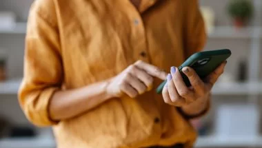 A imagem mostra uma mulher com um celular na mão. O rosto dela não aparece. Ela toca na tela com o dedo indicador, possivelmente em um botão de CTA para realizar uma compra ou outra ação com foco em conversão.