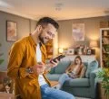 A imagem mostra um homem branco segurando um celular. Ele está fazendo uma compra na sala da casa dele, segurando um cartão de crédito para digitar os dados necessários e concluir a transação.