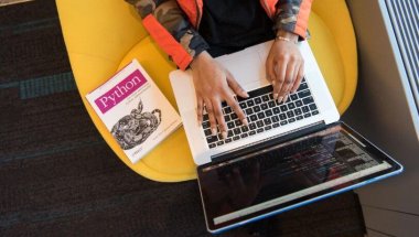 a imagem mostra a visão superior de uma pessoa em um sofá amarelo usando um notebook e ao lado há um livro da linguagem de programação Python.