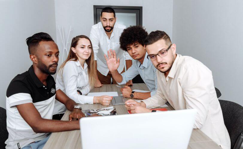 Um grupo de quatro rapazes e uma mulher estão em volta de uma mesa de reunião, interagindo com um notebook. 