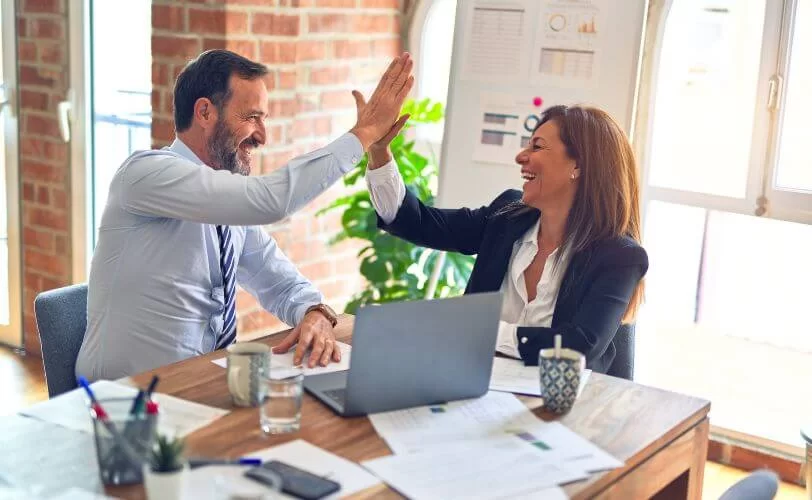 a imagem mostra um executivo e uma executiva felizes em um ambiente semelhante a um escritório. Os dois estão fazendo um high five. A imagem faz referência ao sucesso em comunicação empresarial abordado no conteúdo.