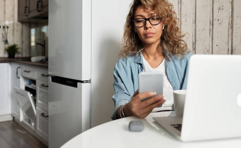 a imagem mostra uma mulher trabalhando em frente a um notebook e ao fundo está uma cozinha. Ela está olhando um smartphone. 