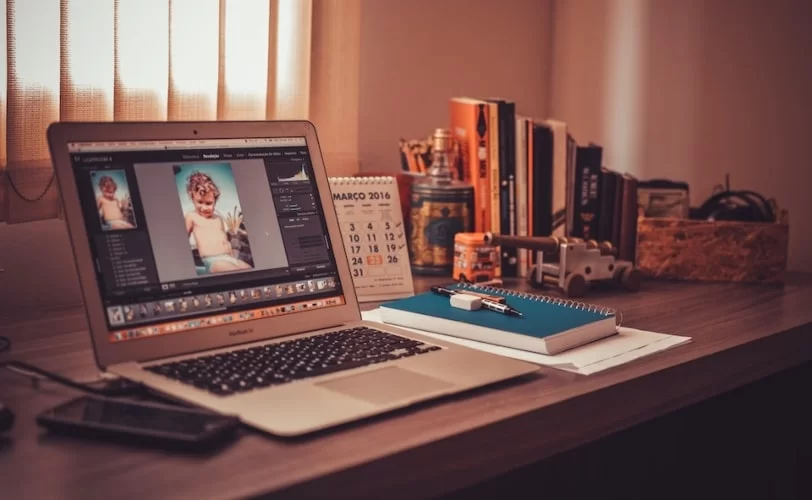 A imagem mostra uma escrivaninha e sobre ela há um notebook, com o Adobe Photoshop aberto, programa que foi escrito em C++.