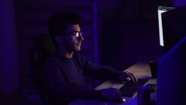 a imagem mostra um rapaz sentado em uma escrivaninha, as luzes do ambiente estão apagadas e a luz das telas de computadores são predominantes. A imagem faz referência a programação na linguagem C++.