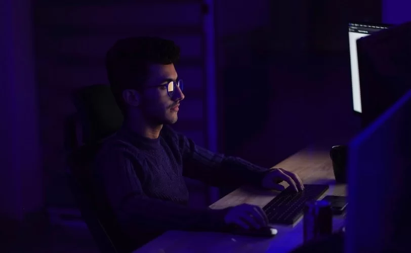 a imagem mostra um rapaz sentado em uma escrivaninha, as luzes do ambiente estão apagadas e a luz das telas de computadores são predominantes. A imagem faz referência a programação na linguagem C++.