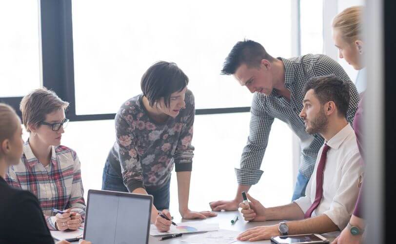 na imagem há uma equipe de seis pessoas, trabalhando em volta de uma mesa. 
