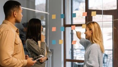 a imagem mostra uma mulher mostrando informações em post-its em uma parede transparente para duas pessoas. A imagem faz referência ao conteúdo sobre o profissional Product Owner.