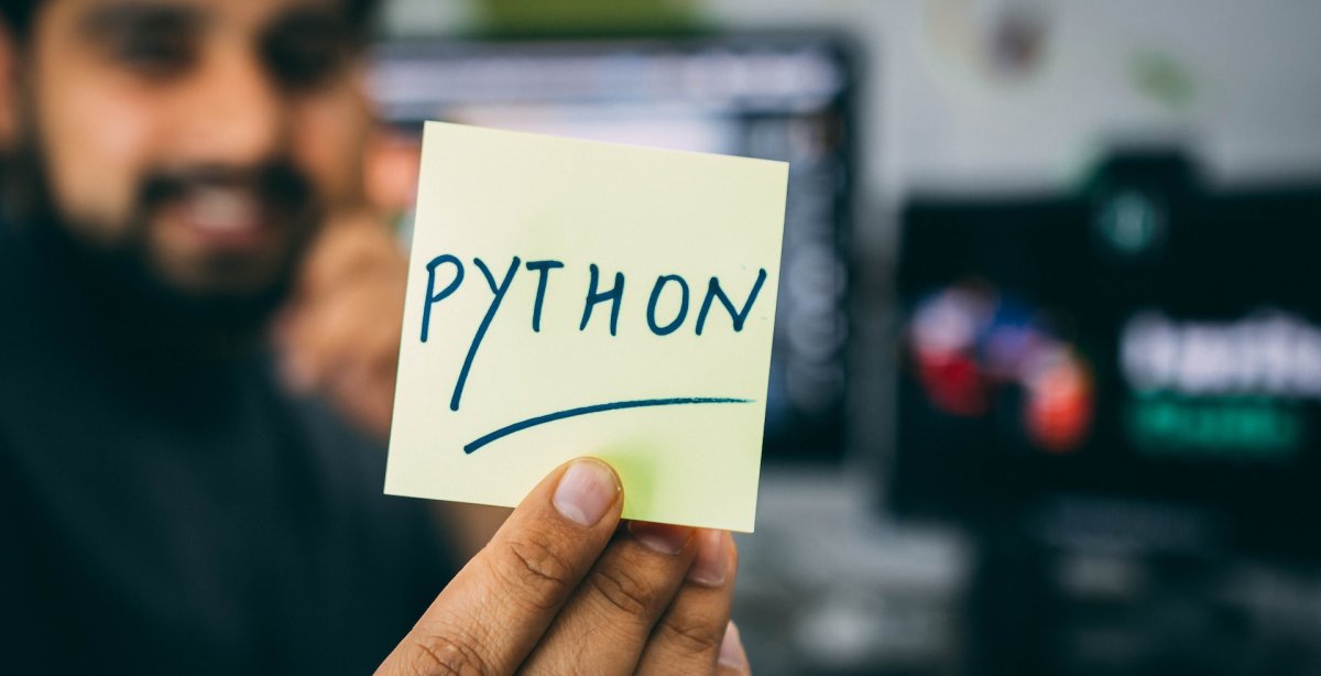 a imagem mostra um homem ao fundo tendo seu rosto desfocado, segurando um bilhete em que está escrito ‘python’ em destaque. A imagem faz referência a biblioteca Matplotlib da linguagem de programação Python.