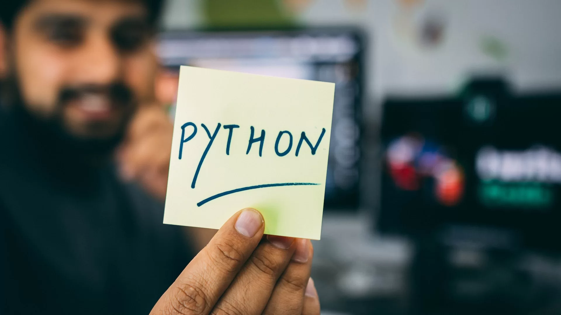a imagem mostra um homem ao fundo tendo seu rosto desfocado, segurando um bilhete em que está escrito ‘python’ em destaque. A imagem faz referência a biblioteca Matplotlib da linguagem de programação Python.