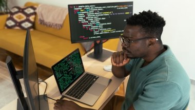 Um jovem desenvolvedor afro-americano está concentrado, analisando um código-fonte em linguagem Go exibido em duas telas de computador. Ele está sentado em uma mesa de trabalho. O ambiente é organizado, com uma decoração aconchegante ao fundo.
