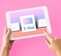 A imagem possui um fundo rosa e há uma pessoa, em que apenas os antebraços estão visíveis, manuseando um tablet. Na tela há a palavra e-mail. A imagem compõe o conteúdo sobre opt-in no email marketing.