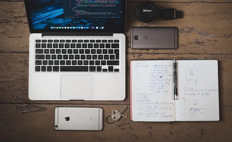 Na imagem temos uma visão de cima de uma mesa de madeira e sobre ela há um notebook, dois celulares, smartwatch, lente de uma câmera nikon e um caderno sem pauta com várias anotações. 