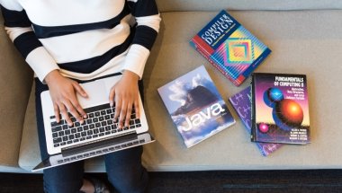 Na imagem temos a visão aérea de uma pessoa sentada em um sofá com uma notebook no colo e ao seu lado há três livros sobre desenvolvimento web.