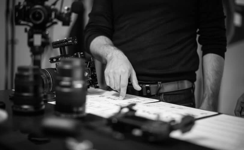 A foto está em preto e branco e há uma mesa com equipamentos de fotografia e aparece o tronco de um homem fazendo apontamento para papéis que estão sobre a mesa. 