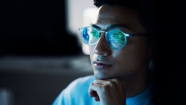 A imagem mostra um homem olhando para uma tela de computador. Ele está programando com o cURL, por isso é possível ver alguns códigos refletidos na cor verde nas lentes de seus óculos.