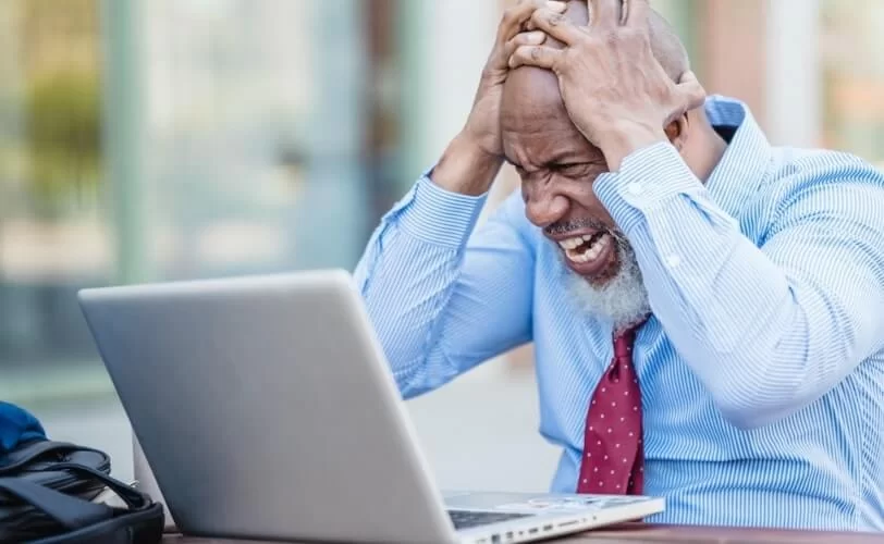 Na imagem há um homem de camisa e gravada com expressão de desespero e as mãos na cabeça. Ele está em frente a um notebook. A imagem faz parte do conteúdo sobre sites fora do ar. 