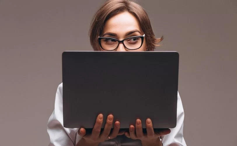 Uma mulher está segurando um notebook. Ela aparece atrás do equipamento, mostrando apenas da metade do rosto para cima.