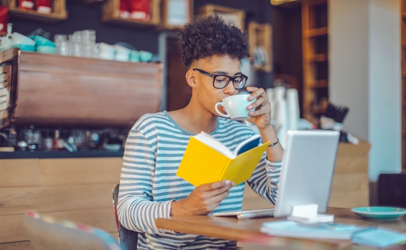 Na imagem, um moça está em um café, lendo um livro e em frente a um notebook. 
