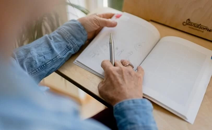 A imagem mostra uma pessoa, no enquadramento aparece as mãos e ombros, e ela está desenhando em um caderno sem pauta. 