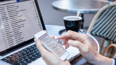 Na imagem aparece apenas as mãos de uma mulher mexendo em um celular e a frente há um notebook e café sobre uma mesa.
