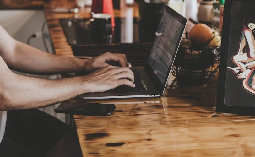 A imagem mostra um homem sentado a uma mesa, digitando em um notebook. 