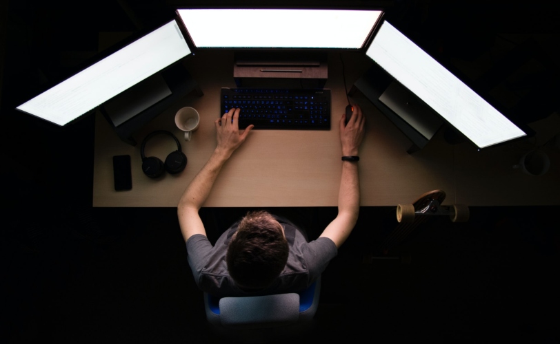Na imagem, um homem está em frente a um computador com três telas.
