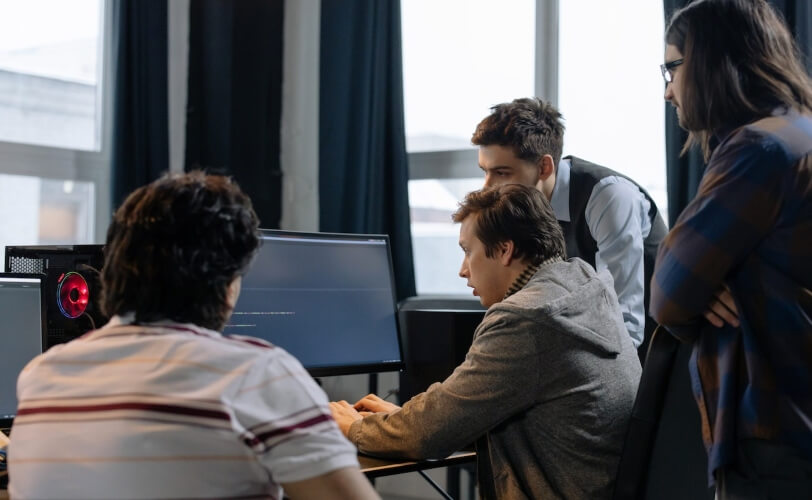 A imagem mostra três homens trabalhando em um escritório em volta de um mesmo computador.