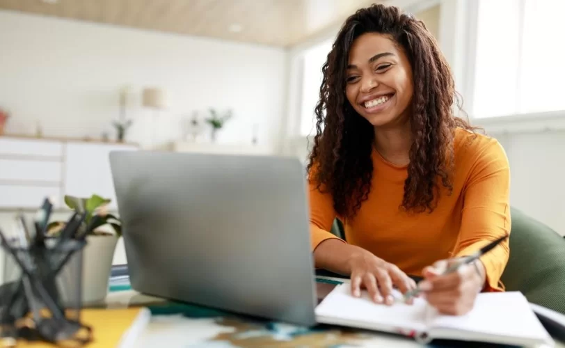 A imagem mostra uma moça sorrindo, olhando para a tela do notebook. Ela também está escrevendo em um caderno. 