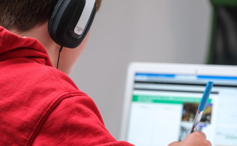 A imagem mostra um rapaz de vermelho escrevendo com uma caneta e olhando para uma tela de notebook. 