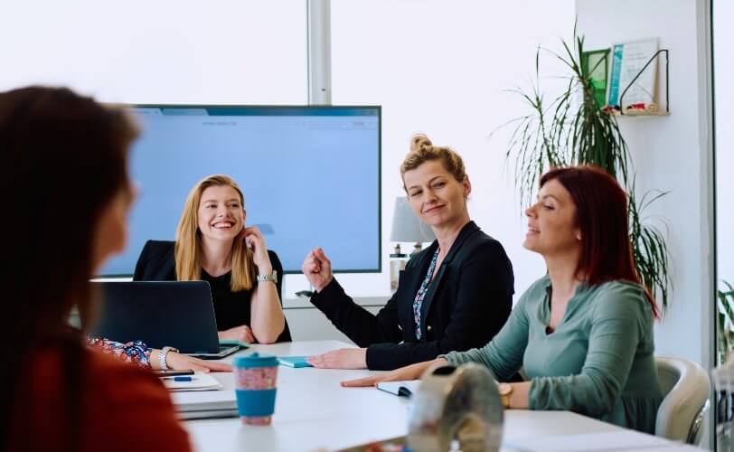 Quatro mulheres conversam sobre um projeto em um escritório. Eles utilizam equipamentos eletrônicos, como notebooks. Atrás do grupo há uma tela de apresentação. A reunião é uma forma de aumentar a produtividade em um time. 