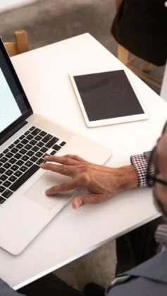 um homem de cabelos escuros, vestindo uma camisa xadrez e um blazer cinza, trabalha em uma apresentação em seu notebook para aumentar sua produtividade. Ele está acomodado em uma pequena mesa branca e tem um tablet sobre ela, ao lado do notebook.