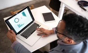 um homem de cabelos escuros, vestindo uma camisa xadrez e um blazer cinza, trabalha em uma apresentação em seu notebook para aumentar sua produtividade. Ele está acomodado em uma pequena mesa branca e tem um tablet sobre ela, ao lado do notebook.