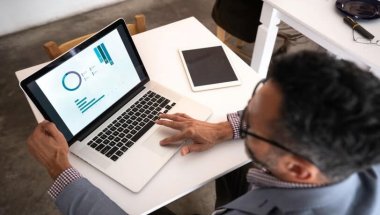 um homem de cabelos escuros, vestindo uma camisa xadrez e um blazer cinza, trabalha em uma apresentação em seu notebook para aumentar sua produtividade. Ele está acomodado em uma pequena mesa branca e tem um tablet sobre ela, ao lado do notebook.