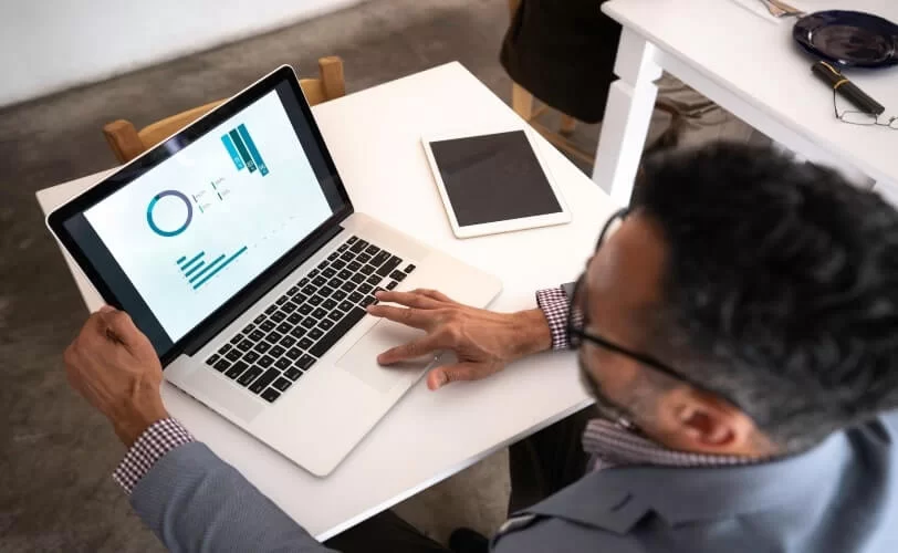 um homem de cabelos escuros, vestindo uma camisa xadrez e um blazer cinza, trabalha em uma apresentação em seu notebook para aumentar sua produtividade. Ele está acomodado em uma pequena mesa branca e tem um tablet sobre ela, ao lado do notebook.