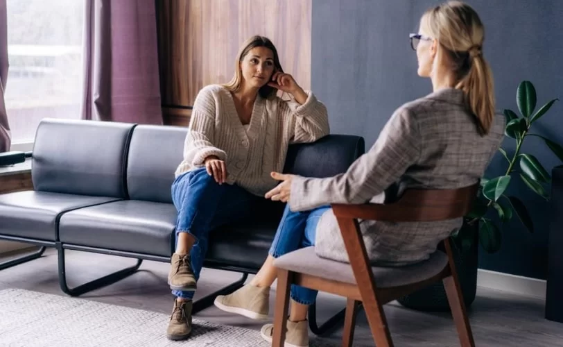 Na imagem estão duas mulheres sentadas uma frente a outra conversando. 