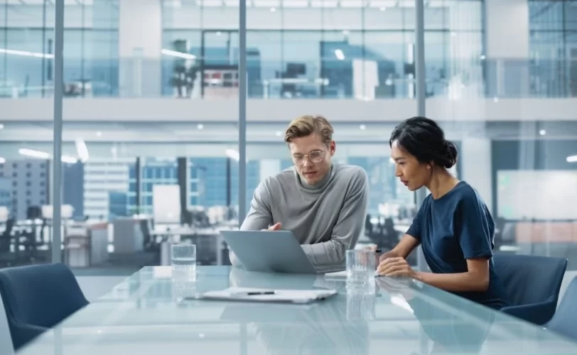 Na imagem um homem e mulher estão sentados em uma mesa de reunião e ambos estão prestando atenção em um notebook.